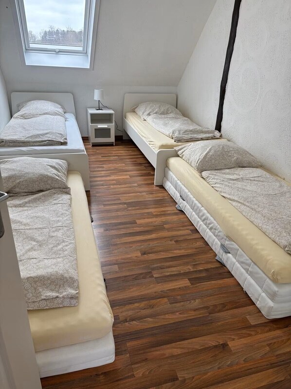 Een kleine, compacte kamer met drie bedden op een rij, een witte nachtkastje en een dakvenster erboven.