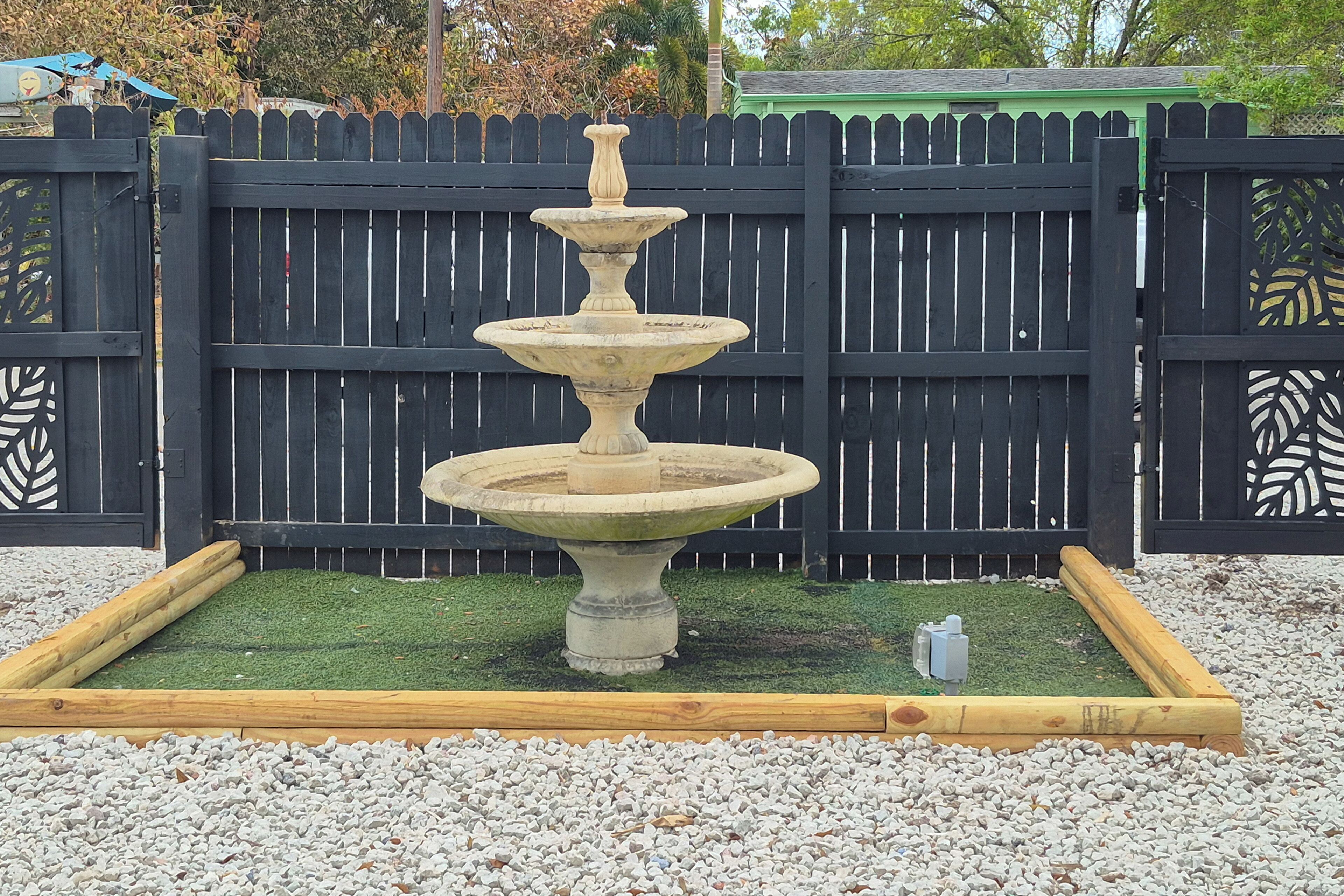 Une fontaine en pierre à plusieurs niveaux, située dans un jardin bordé de bois, avec une clôture en piquets noirs en arrière-plan.