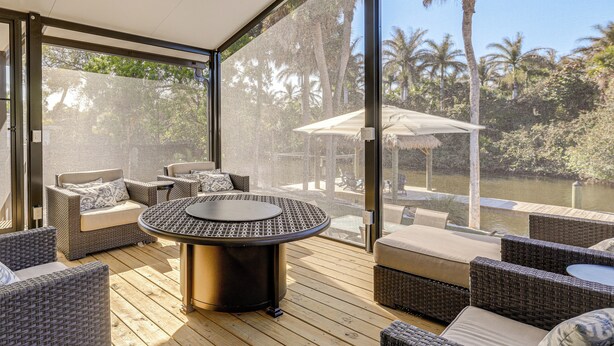 A modern outdoor patio with wicker furniture, a round table, and a glass-enclosed area offering a view of palm trees and a water body. - Riverfront Oasis: Fish, Grill & Chill in Stuart! (Stuart)