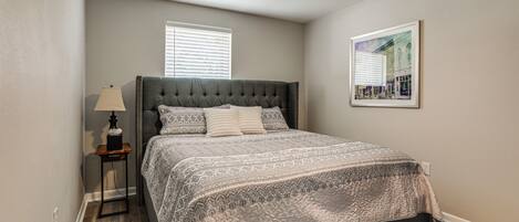 A bedroom with a tufted headboard, a patterned bedspread, a bedside lamp, and a ceiling fan.
