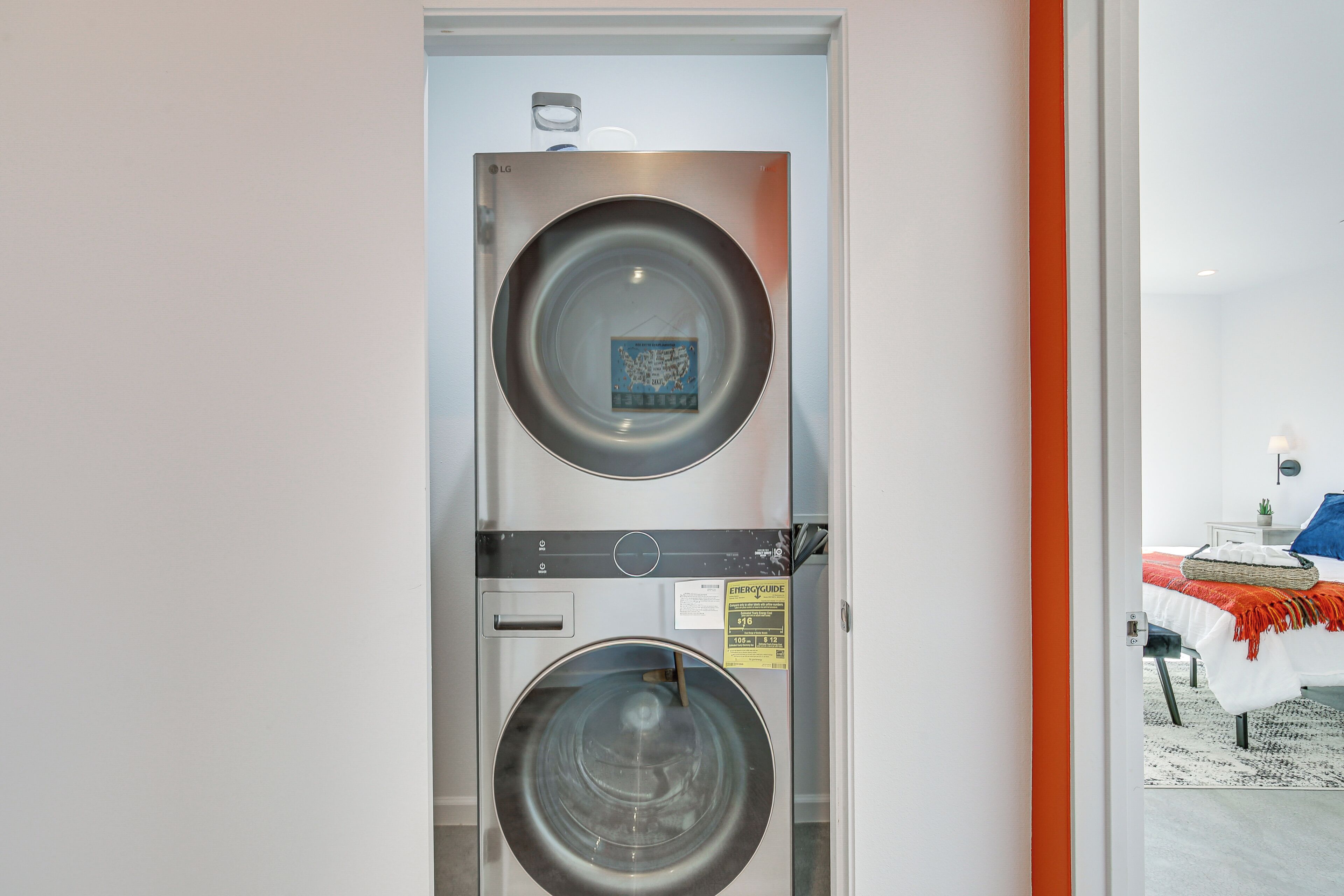 Ein Waschraum mit zwei gestapelten Waschmaschinen, einem sichtbaren Energieeffizienzlabel und im angrenzenden Raum ein Bett mit roter Decke.