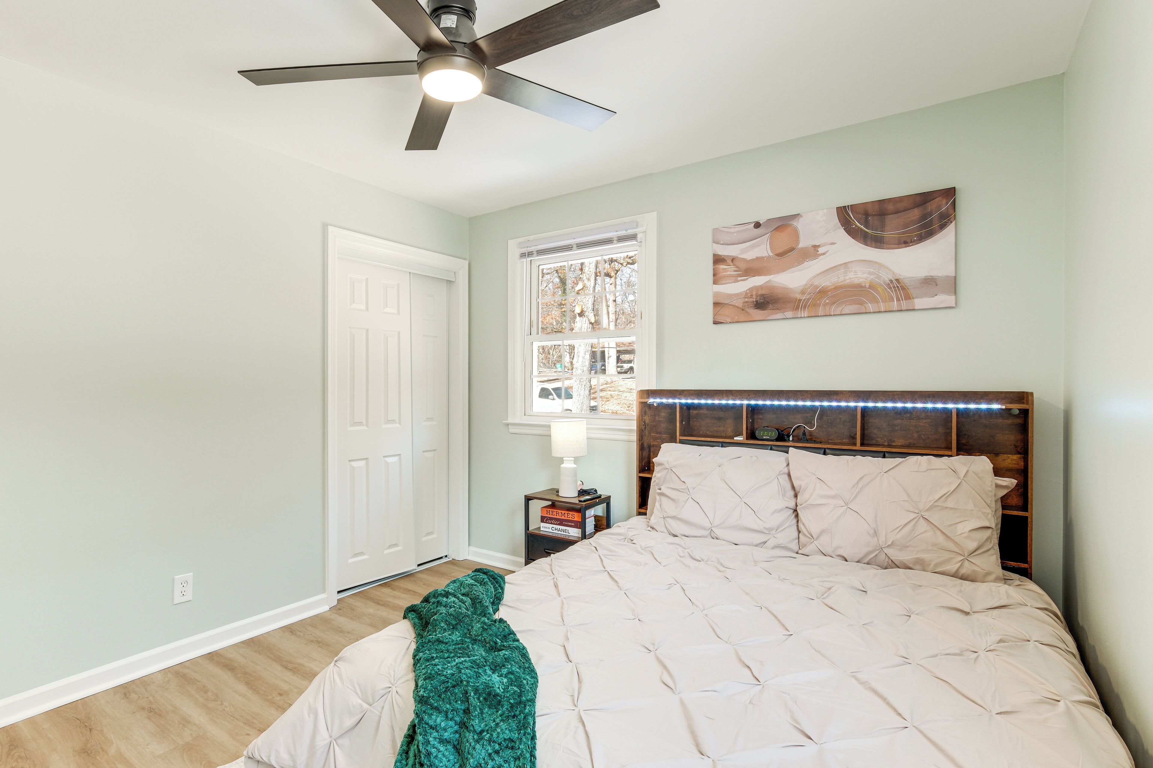 A bedroom with a bed, a ceiling fan, a nightstand, a lamp, and a window with a view of trees.
