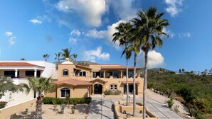 A large, multi-story house with a spacious patio area, surrounded by palm trees and cacti. - Steps to Sand: Ocean-view Cabo Home w/ Heated Pool (Cabo San Lucas)