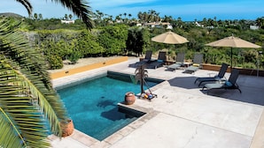 A pool area with lounge chairs and umbrellas, surrounded by greenery and a view of the ocean. - Steps to Sand: Ocean-view Cabo Home w/ Heated Pool (Cabo San Lucas)