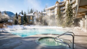 A hot tub surrounded by a pool, with a building featuring multiple balconies and a snowy landscape in the background. - Whistler - Cascade Lodge 117 (Whistler)