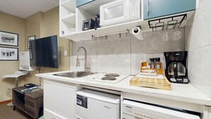 Een moderne keuken met een spoelbak, fornuis, magnetron en koffiezetapparaat. Er is een kleine koelkast, een afwasschuur en een houten snijplank met een mes.