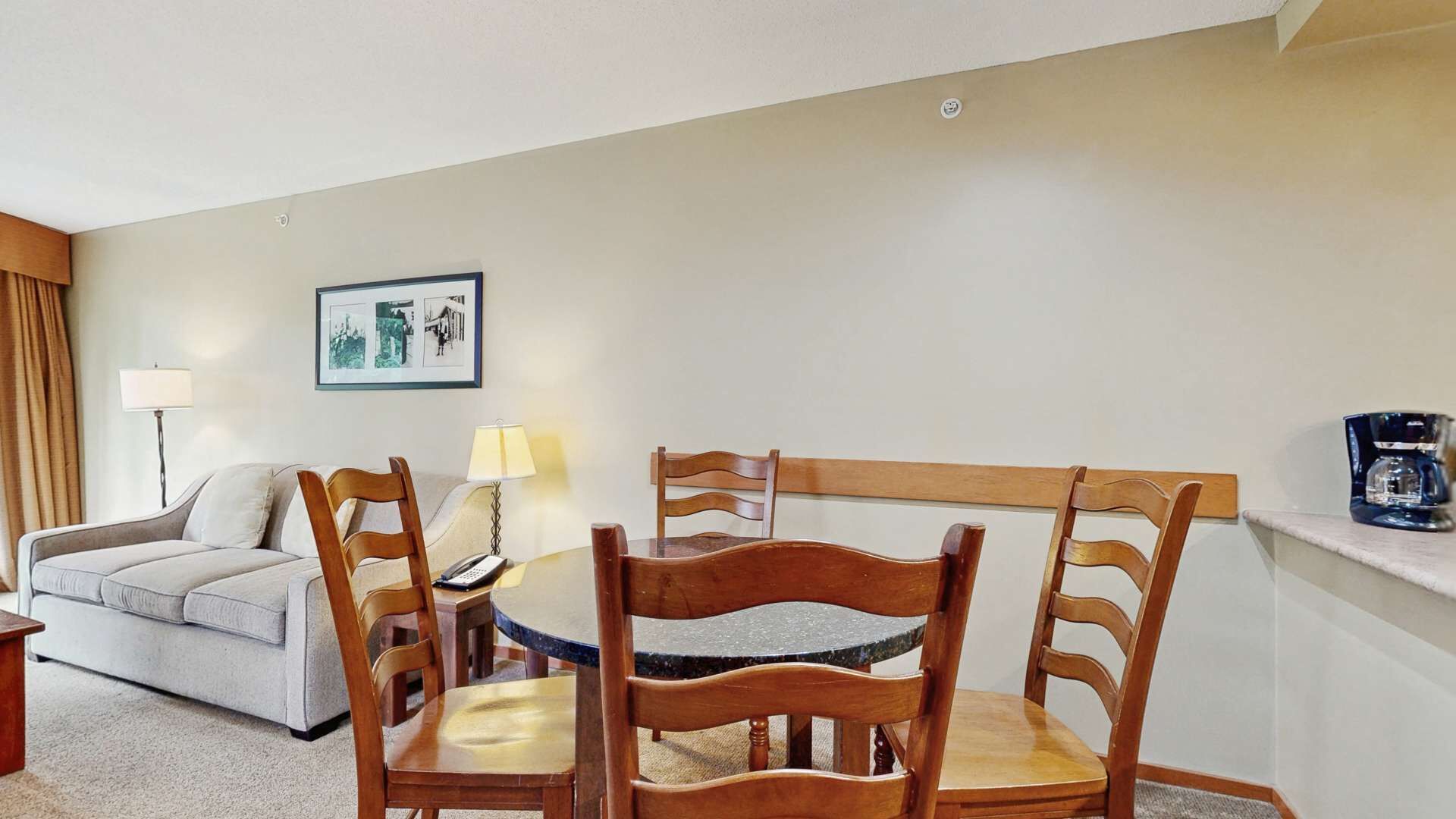 Ein Hotelzimmer mit einem runden Esstisch, vier Holzsesseln, einer Couch, einer Lampe, einem gerahmten Bild an der Wand und einer Kaffeemaschine auf einem Regal.