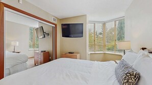 Quarto de hotel com uma cama grande, uma televisão de tela plana na parede, uma escrivaninha com luminária e uma janela com persianas.