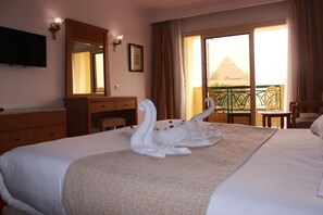 A hotel room with a bed, a desk, a chair, a TV, a balcony with a view of a pyramid. - Regency Pyramids Hotel (Giza)