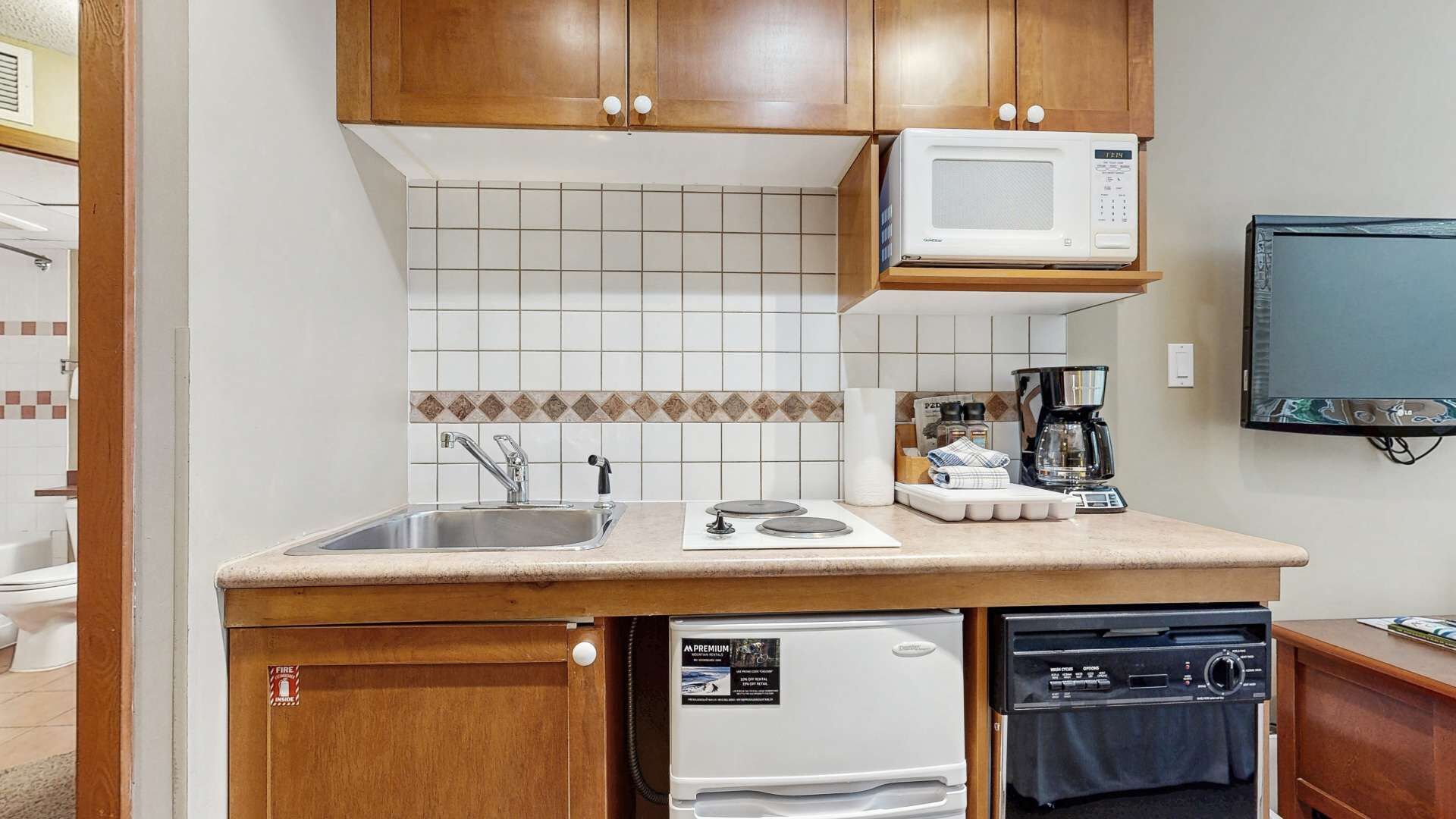 A compact kitchen with wooden cabinets, a microwave, a coffee maker, a dishwasher, and a television on the wall.
