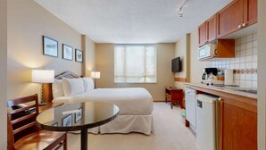 Ein Hotelzimmer mit Bett, Nachttisch, Lampe, Sessel, rundem Tisch, einer Küchenzeile und einem Fernseher.