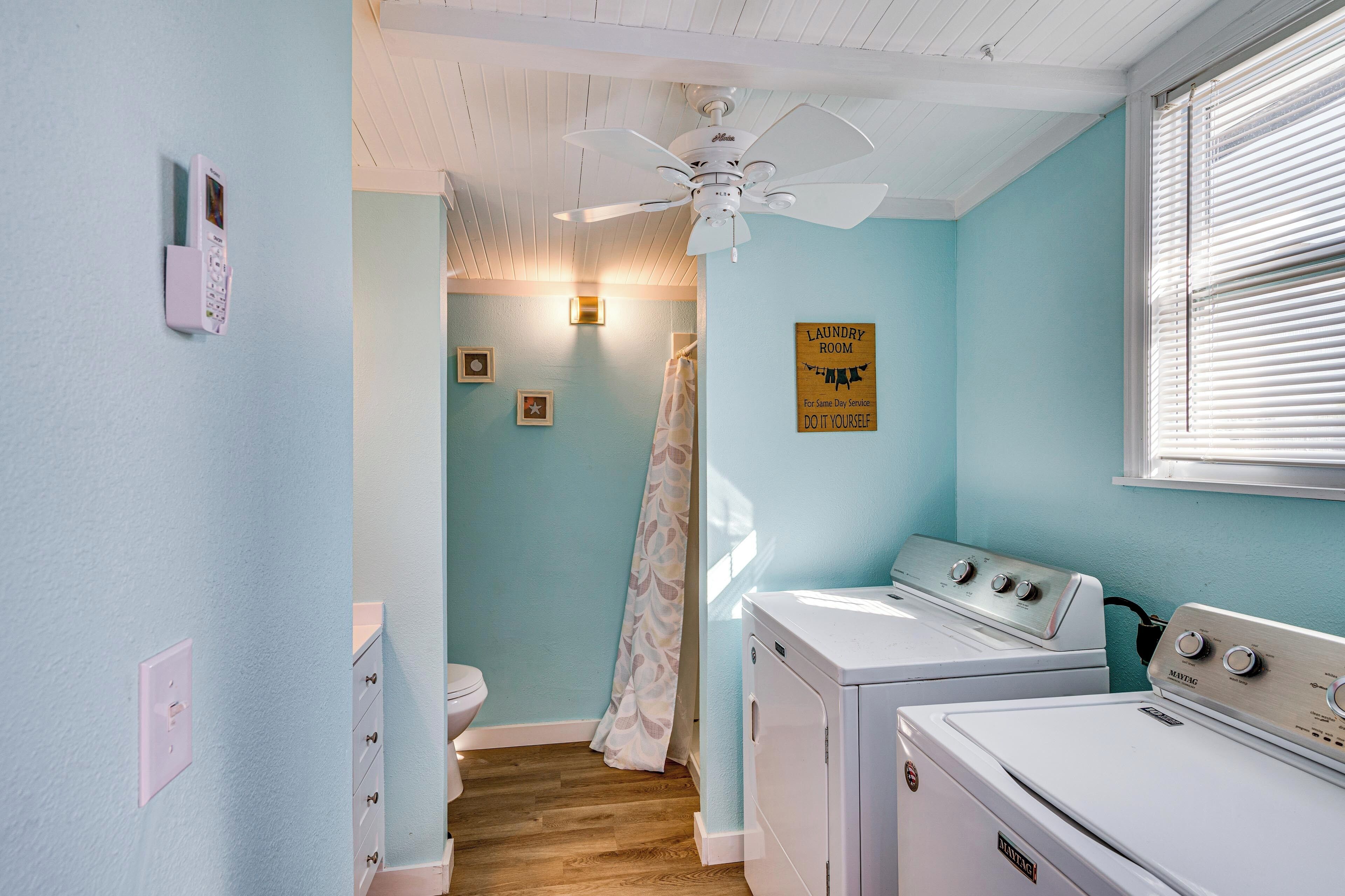 A laundry room with two white front-loading washers and dryers, a ceiling fan, and a sign indicating the room's purpose.