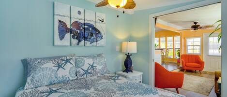 A bedroom with a bed, a ceiling fan, artwork, and a view into another room.