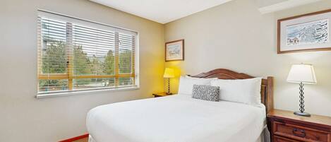 Una habitación de hotel con una cama grande, una mesita de noche con lámpara, una ventana con persianas y un cuadro en la pared.