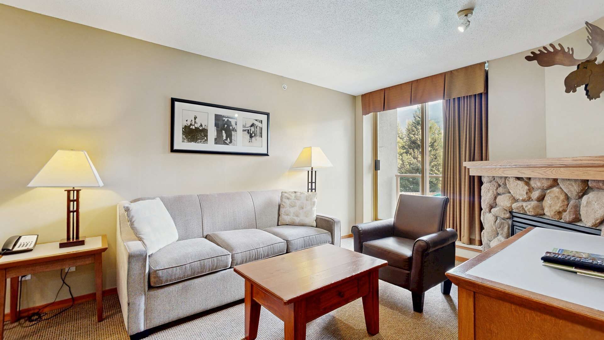 A hotel room with a grey sofa, a brown leather chair, a wooden coffee table, and a fireplace. There are two lamps on side tables and a framed picture on the wall.