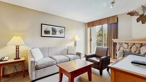 A hotel room with a grey sofa, a brown leather chair, a wooden coffee table, and a fireplace. There are two lamps on side tables and a framed picture on the wall. - Whistler - Cascade Lodge 212 (Whistler)