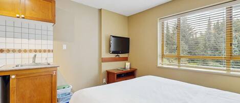 Une chambre d’hôtel avec un lit, une petite kitchenette, une télévision et une fenêtre avec des stores.