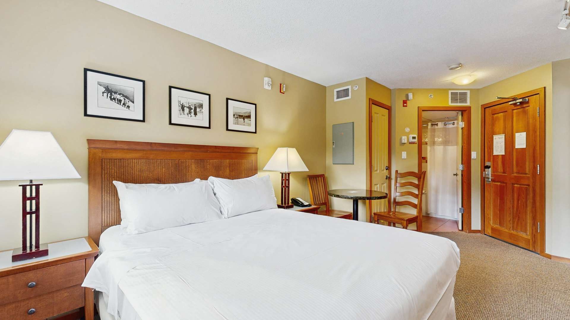 Une chambre d’hôtel avec un grand lit, deux tables de chevet avec des lampes, une petite table ronde avec une chaise, et des tableaux encadrés au mur.