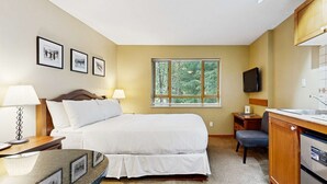 A bedroom with a bed, bedside lamps, a desk, a chair, a window with blinds, a TV, and a kitchenette. - Whistler - Cascade Lodge 225 (Whistler)