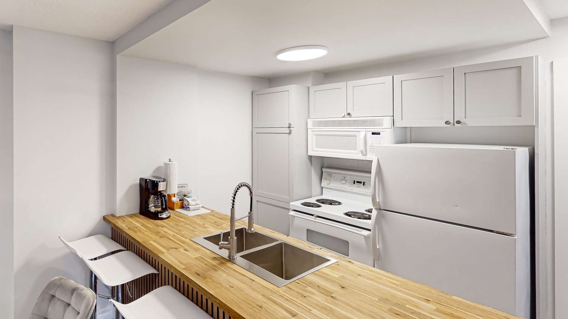 Cocina compacta con armarios blancos, encimera de madera, lavamanos, cafetera, microondas y refrigerador.