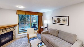 A living room with a stone fireplace, a sofa, a chair, a coffee table, and a large window with a view of a backyard. - Whistler - Cascade Lodge 232 (Whistler)