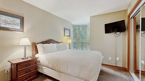 Une chambre à coucher comprenant un lit, une table de chevet, une lampe, un téléviseur fixé au mur et une fenêtre avec des stores.