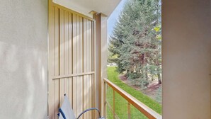 Un balcon avec un volet en bois, une chaise en métal et une vue sur un jardin avec des arbres et de l’herbe.