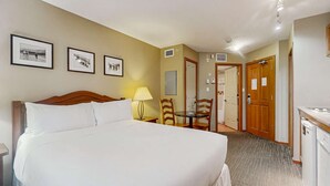 Habitación de hotel con una cama grande, un escritorio con lámpara, una silla y una mesa de comedor. Se observan cuadros enmarcados en la pared y una puerta que da a otra habitación.
