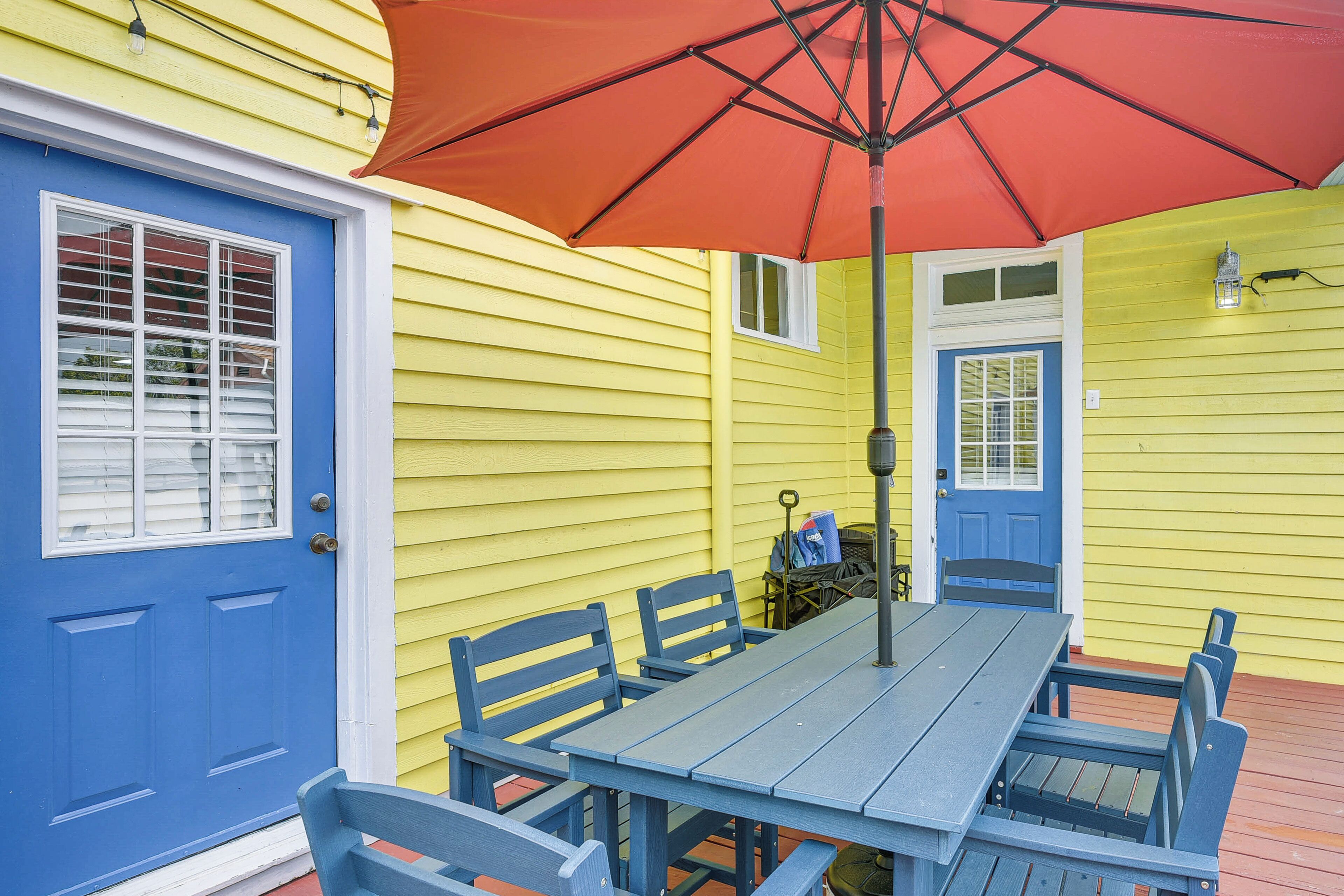 Une table bleue, assortie à des chaises, et un parasol rouge, le tout situé devant un bâtiment jaune aux portes et fenêtres bleues.