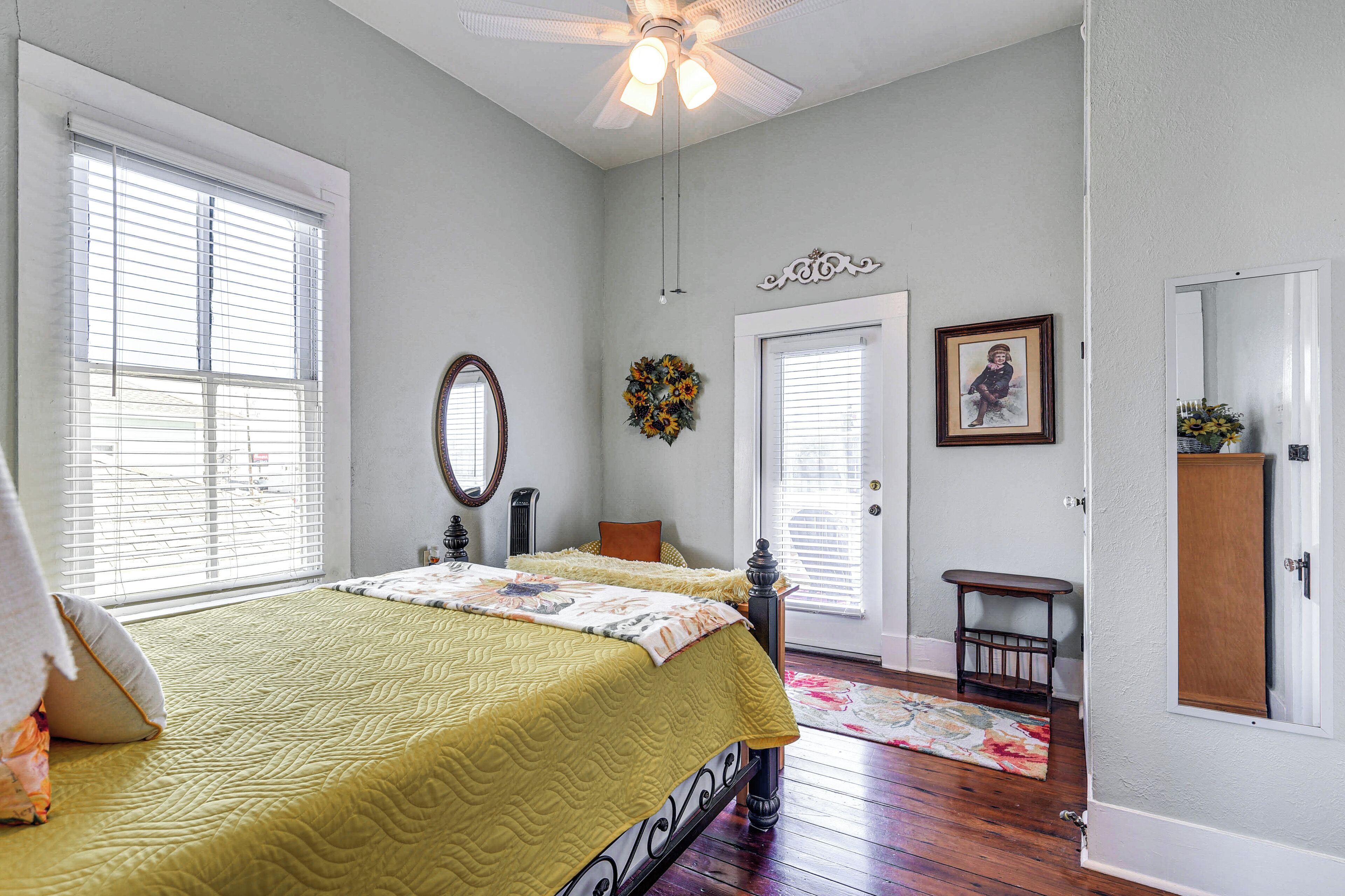 A bedroom with a bed, a ceiling fan, a door, a mirror, and a small table.