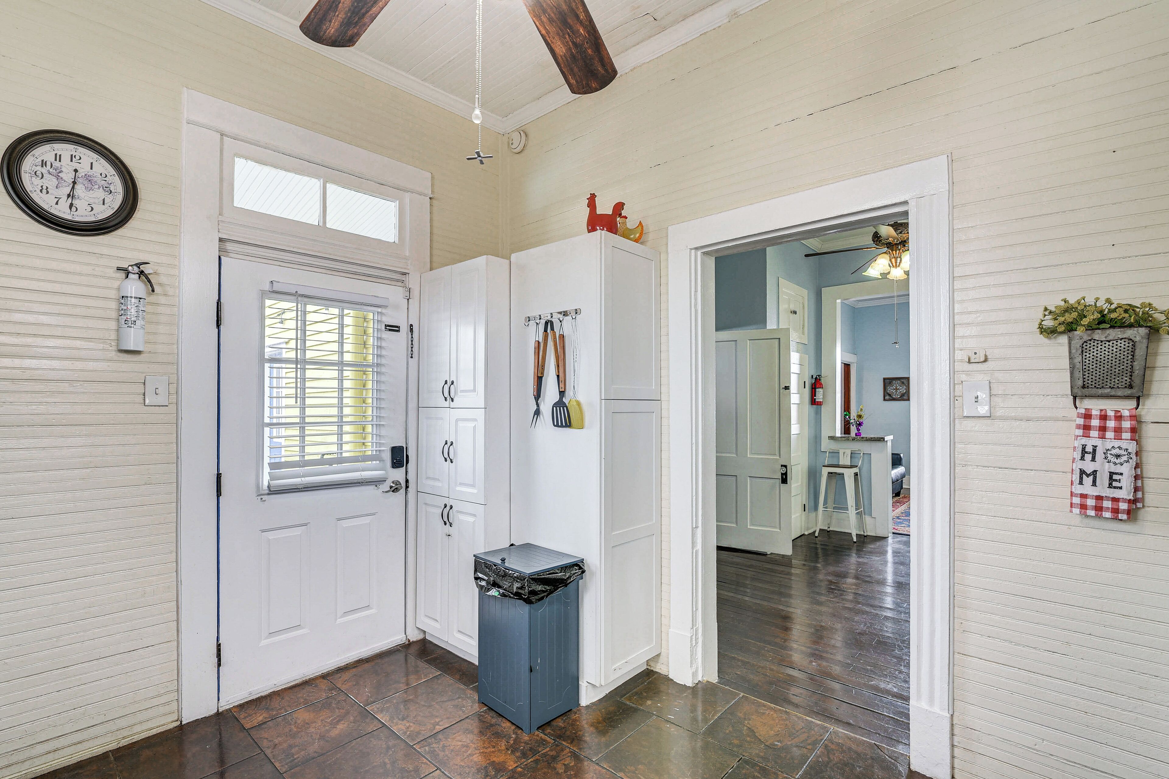 A hallway with a white door, a window with blinds, a trash bin, and a ceiling fan.