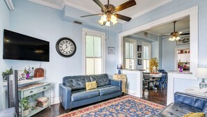 A living room with a blue sofa, a patterned rug, a flat-screen TV, a ceiling fan, and a dining area visible through an open doorway. - Steps to Pleasure Pier: Family Escape w/ Pool! (Galveston)