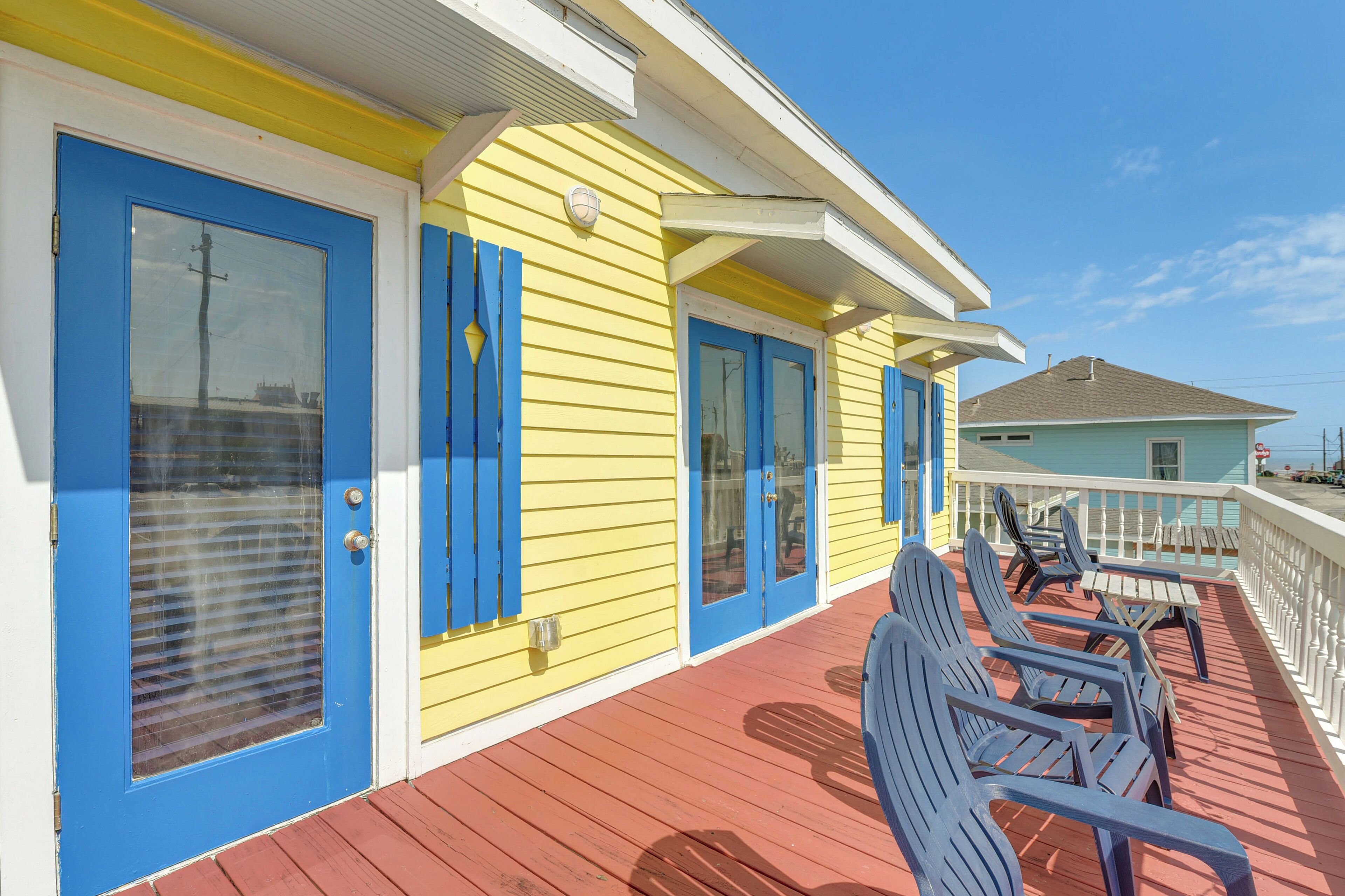A yellow building with blue doors and windows, a wooden deck with chairs, and a white railing.