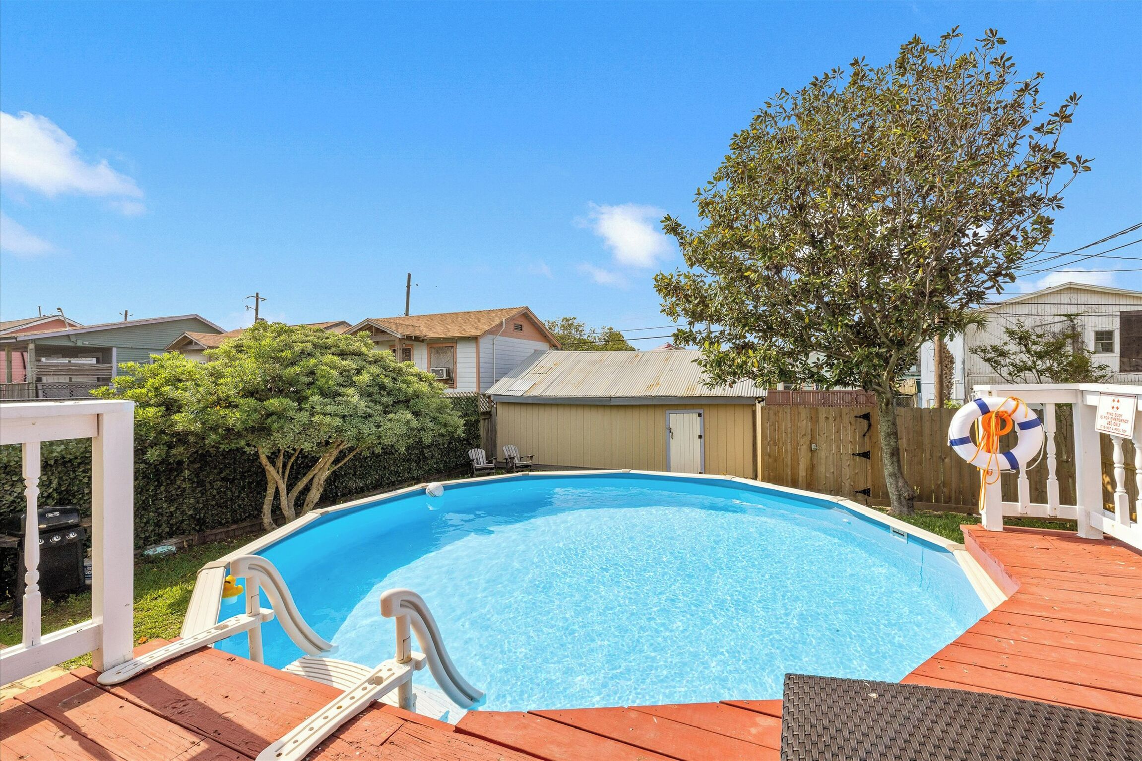 A rectangular swimming pool with a wooden deck, surrounded by a wooden fence, and a tree to the right.
