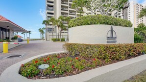 A landscaped area with a circular flower bed, a building with a red roof, and a tall residential structure in the background. - Walk to Sand Key! 11th-floor Clearwater Beach Gem (Clearwater Beach)