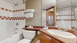 Une salle de bain avec une baignoire, des toilettes et un lavabo. On y trouve un porte-serviettes avec des serviettes, un miroir et une porte qui donne sur une autre pièce.