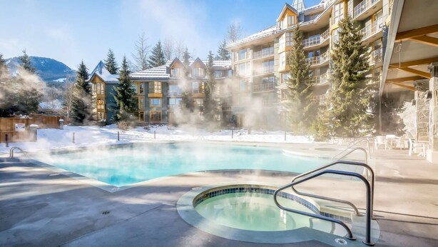 Un bain à remous entouré d’une piscine, avec vue sur un immeuble à plusieurs étages et un paysage enneigé.
