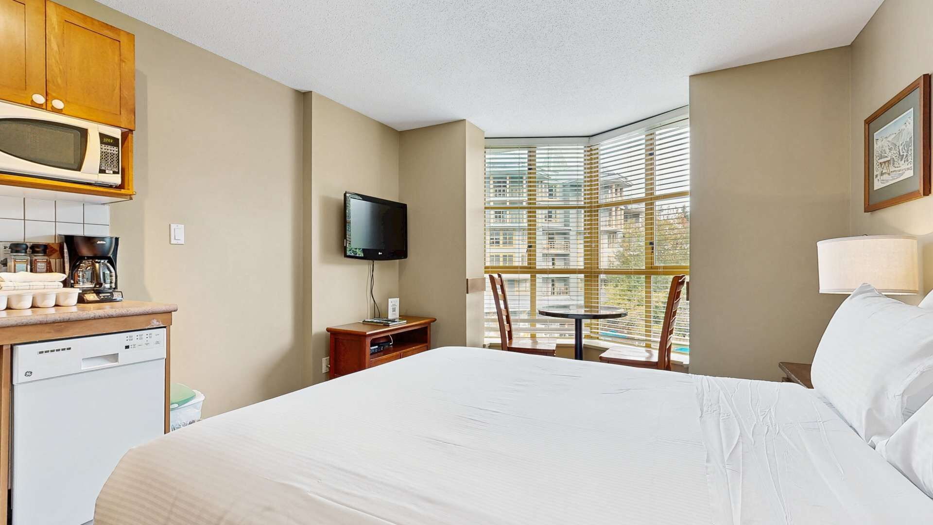 Habitación de hotel con cama, televisor, una mesita con sillas, microondas, cafetera y ventana con persianas.