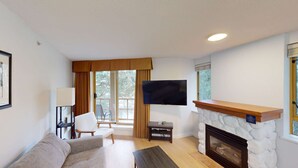 Ein Wohnzimmer mit Kamin, einer Couch, einem Sessel, einem an der Wand montierten Fernseher und einem Fenster mit Vorhängen.