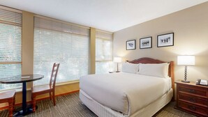 Habitación de hotel con una cama grande, dos lámparas de noche, una mesita de noche de madera, una pequeña mesa con una silla y tres cuadros enmarcados en la pared.