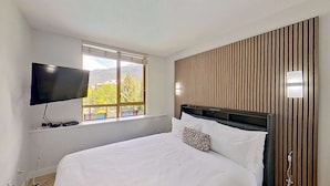 Une chambre d’hôtel avec un lit, un téléviseur fixé au mur, une fenêtre donnant sur des arbres et des montagnes, et un mur recouvert de panneaux en bois.