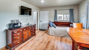 Een kamer met een houten vloer, een aan de muur bevestigde televisie, een houten dressoir, een bed met hoofdeinde en een raam met gordijnen.