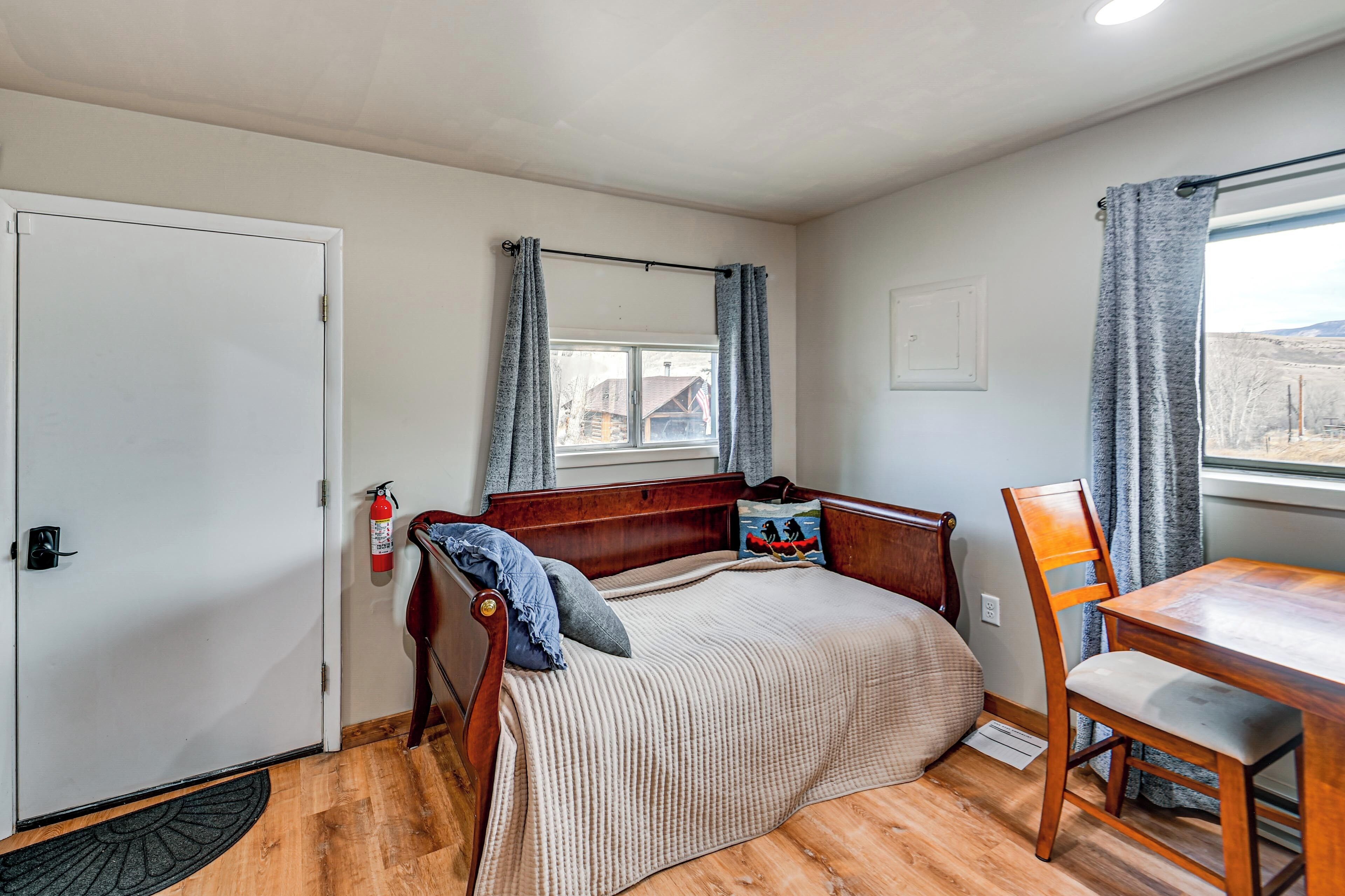 A bedroom with a wooden bed, a wooden chair, a small table, a window with curtains, and a door.