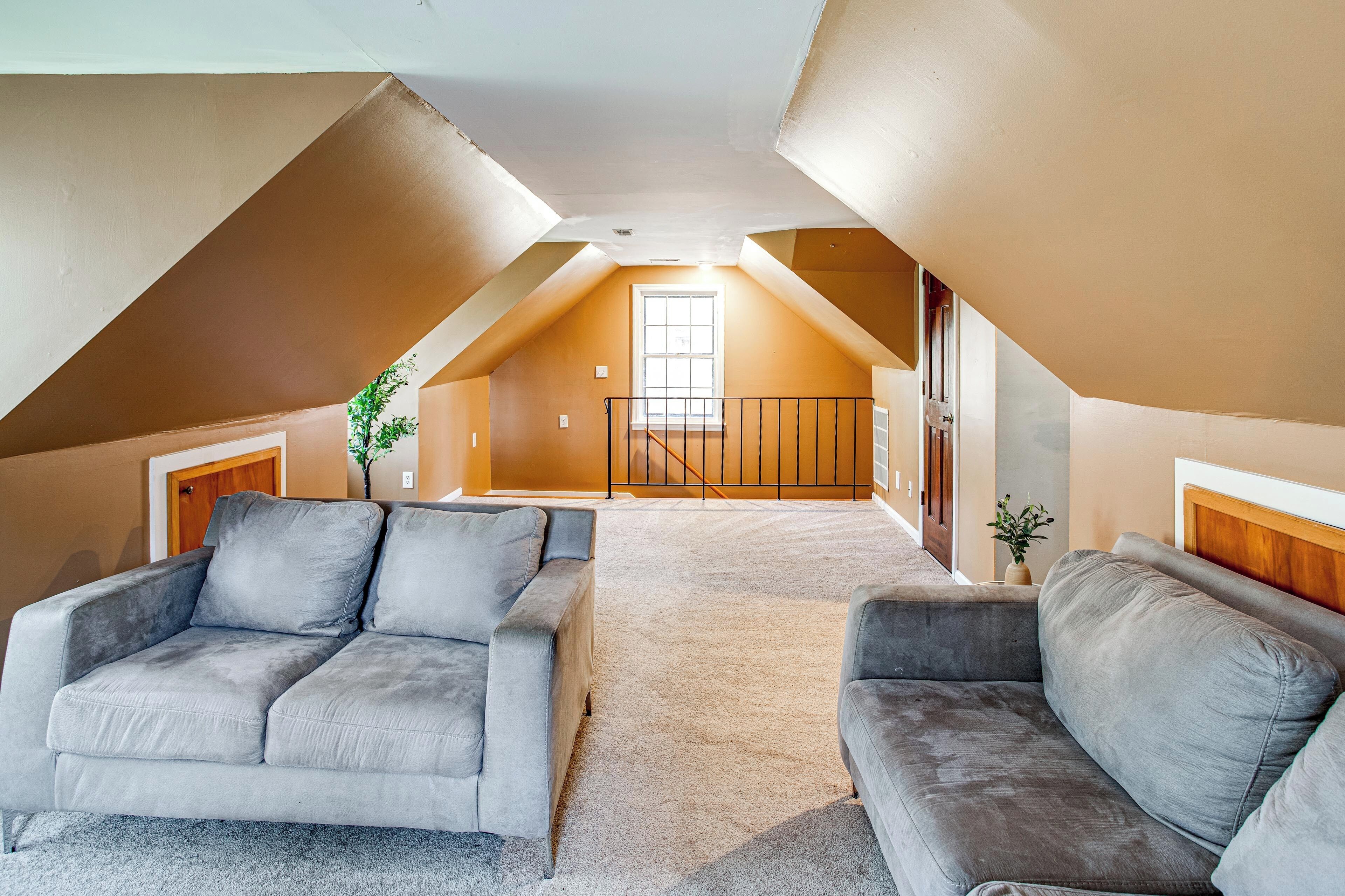 An attic bedroom with a sloped ceiling, two grey sofas, a large window, and a staircase leading to the upper floor.