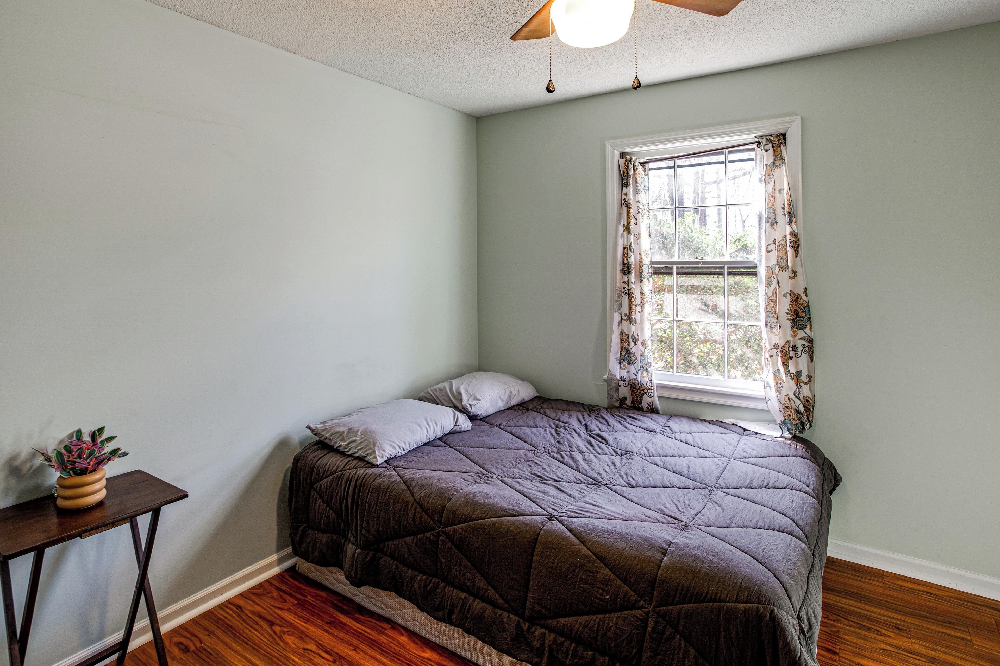 A bedroom with a bed, a window with curtains, a ceiling fan, and a small table with a plant.