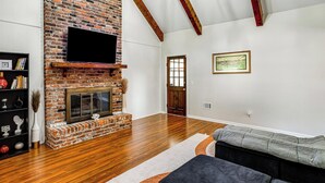 A living room with a brick fireplace, wooden beams, a flat-screen TV, a bookshelf, and a sofa. - Lake Murray Retreat Near USC & Columbia (Columbia)