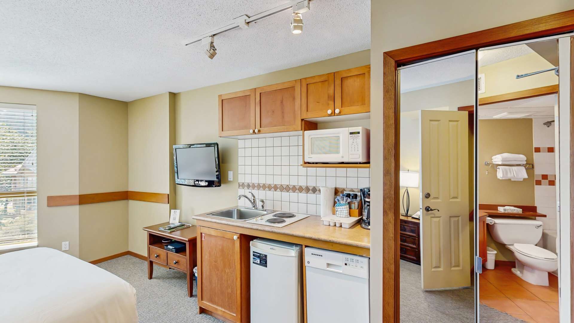 Una habitación de hotel compacta con kitchenette, una cama, televisor y baño con inodoro y ducha.