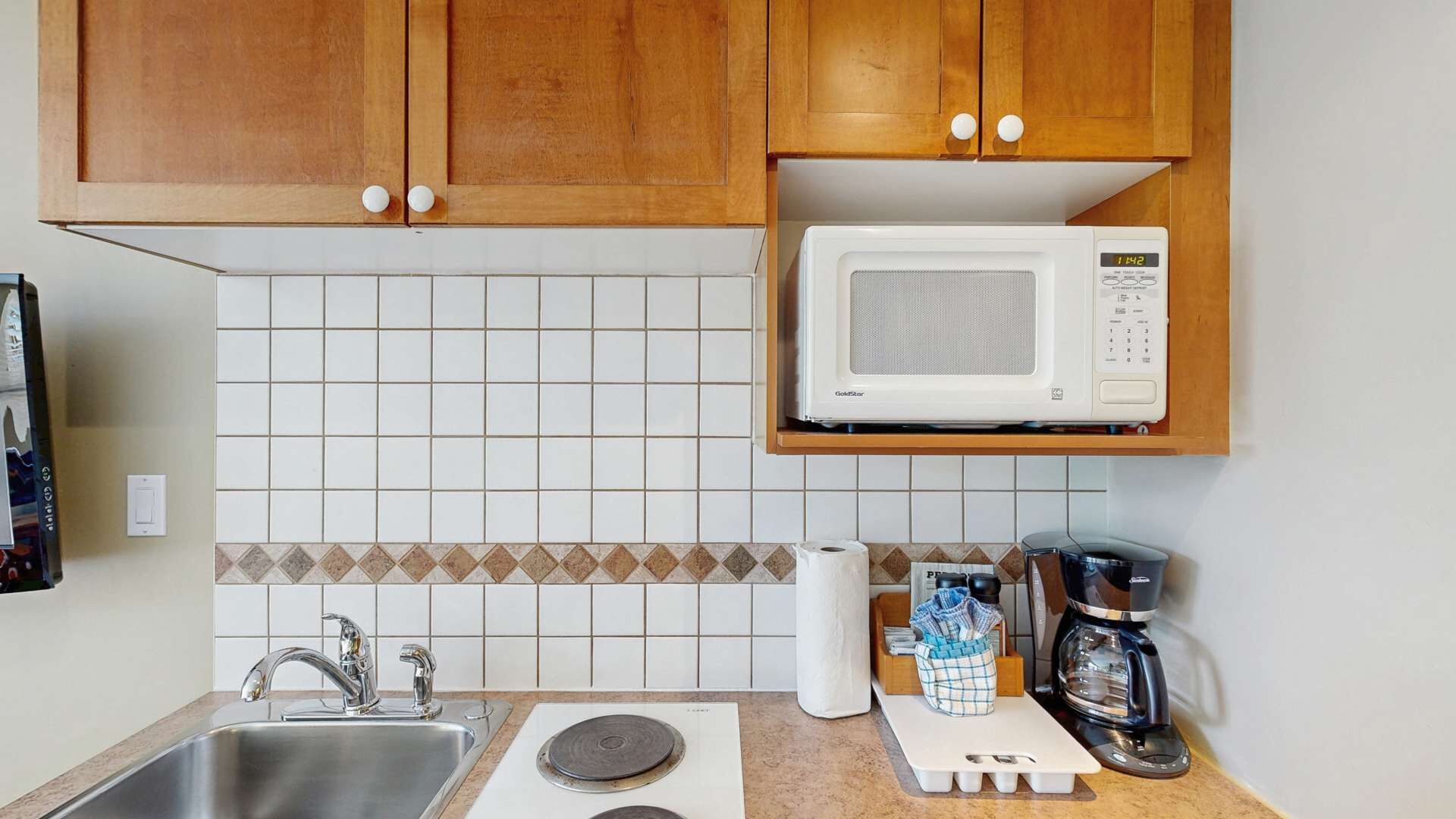 Una cocina con un lavaplatos de acero inoxidable, un microondas, una cafetera y un salpicadero de azulejos.
