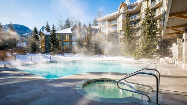 Un jacuzzi rodeado por una piscina, con un edificio que tiene balcones y un paisaje de montañas al fondo.