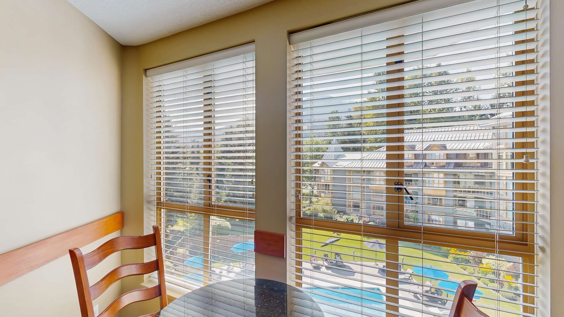Ein Balkon mit Tisch und Stühlen, große Fenster mit Jalousien und Blick auf ein Schwimmbad und Gebäude.